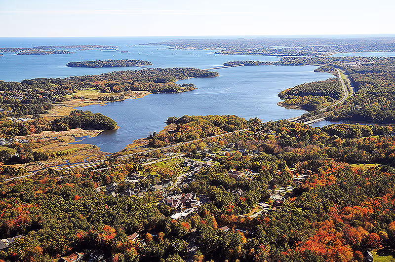 Aerial view of a large body of water surrounded by dense forest with autumn foliage. A road runs alongside the water, and a small community or facility is visible among the trees near the shoreline.