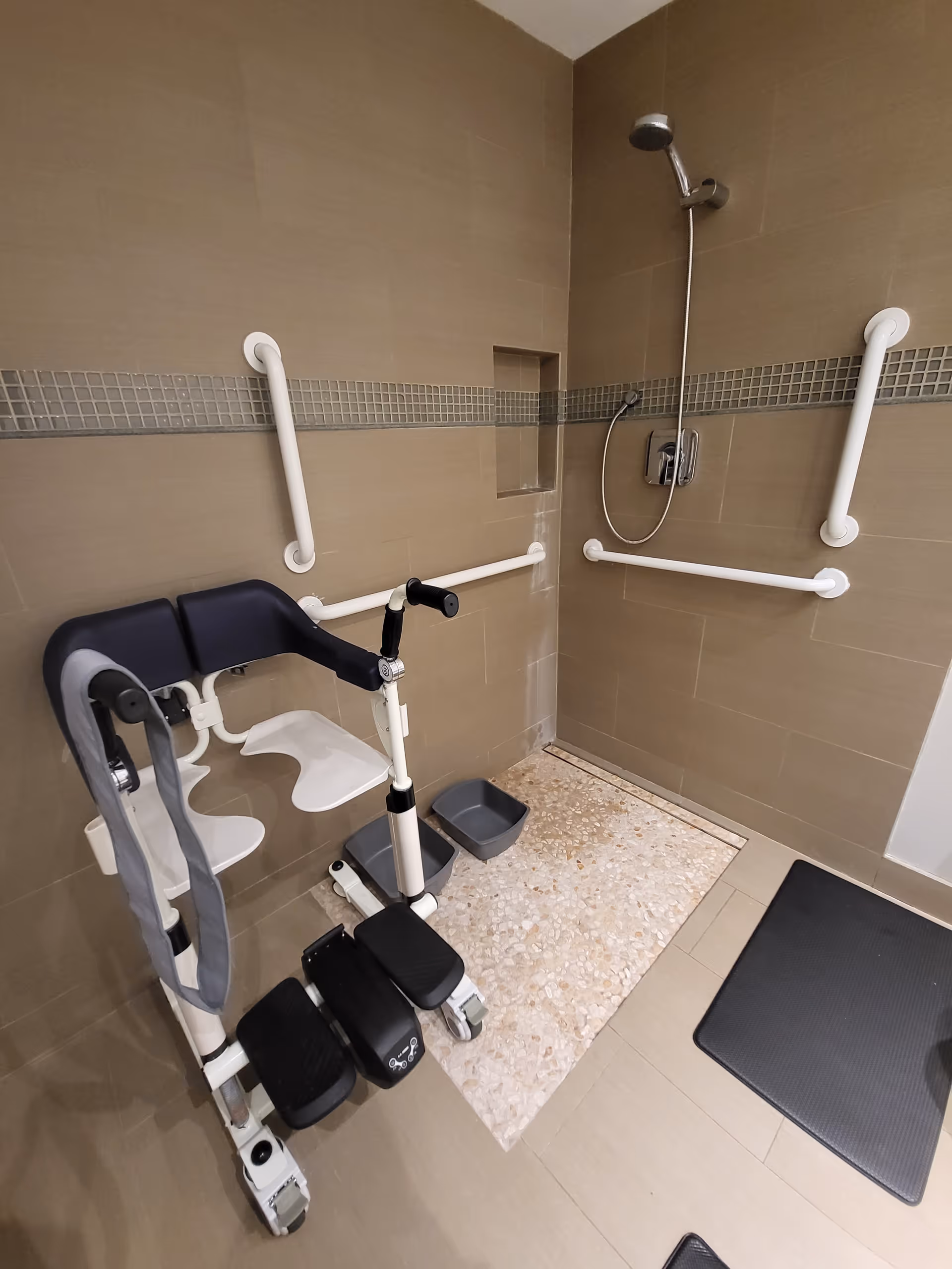Accessible shower area with beige tiled walls and floor, equipped with white grab bars on the walls. There is a showerhead attached to the wall and a specialized shower chair with footrests and armrests positioned inside the shower space.