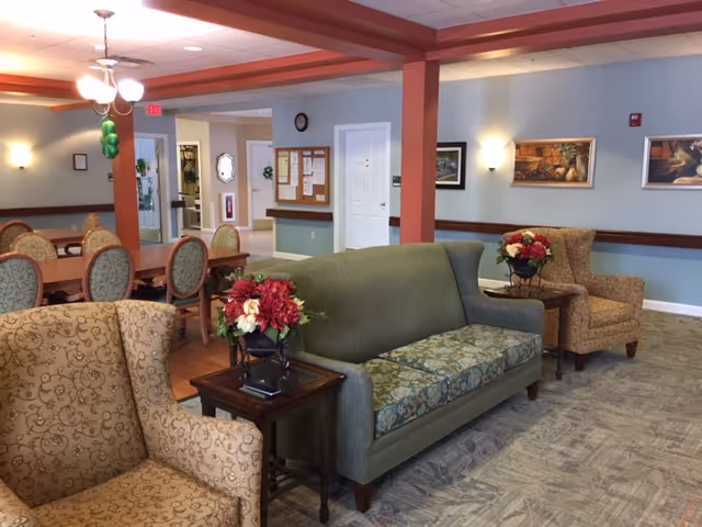 A cozy common area in a senior living facility featuring a green upholstered sofa with floral cushions, two patterned armchairs, and wooden side tables with flower arrangements. In the background, there is a dining area with a wooden table and chairs, light blue walls adorned with framed artwork, and a bulletin board near a white door.