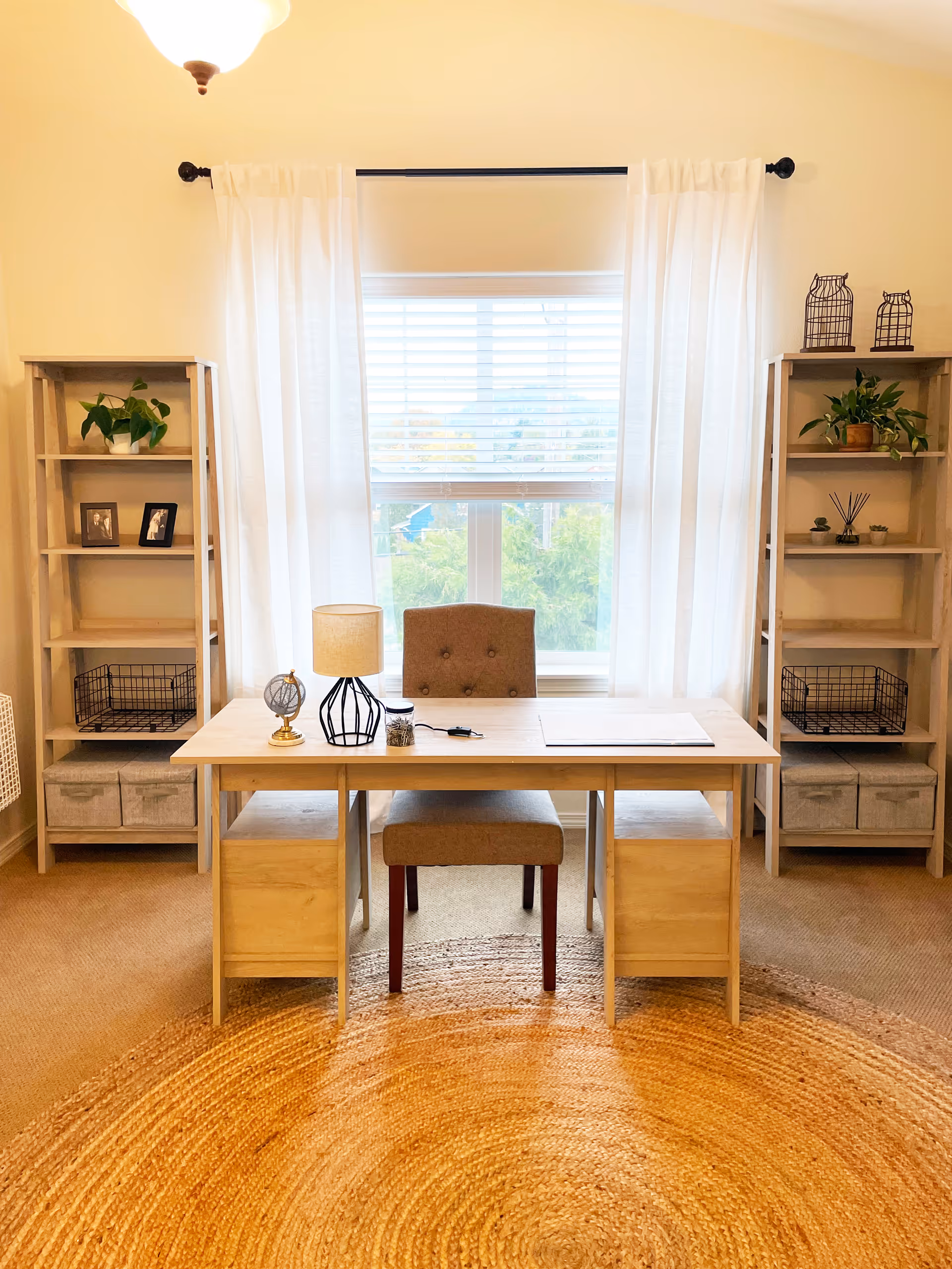 A bright room with a wooden desk and a cushioned chair in front of a window with white curtains and blinds. On the desk are a small lamp, a decorative globe, and some small items. On either side of the window are matching wooden shelves with plants, framed photos, and decorative items. The floor has a large round woven rug.
