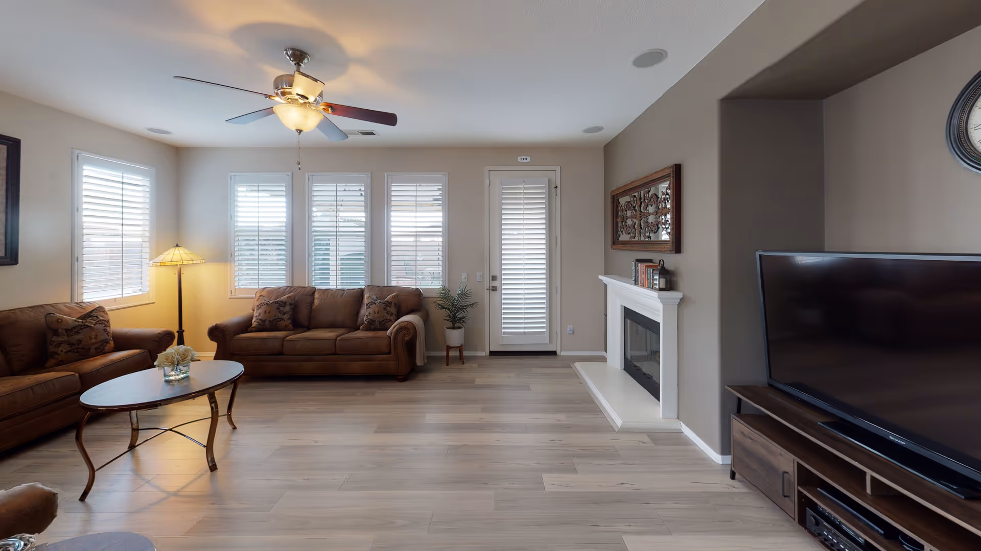 A well-lit living room with two brown sofas, a wooden coffee table with a flower vase, a floor lamp, a ceiling fan with lights, a TV on a wooden stand, a fireplace with decorative items on the mantel, and windows with white shutters.