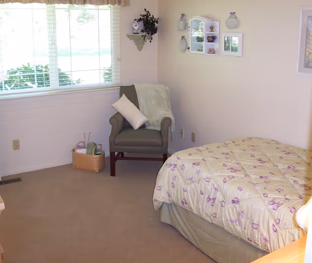 A cozy bedroom with a bed covered in a floral patterned comforter, a green armchair with a white pillow and a light green throw blanket, a basket with towels and toiletries on the floor, and a window with blinds letting in natural light. The walls are decorated with small shelves and framed pictures.