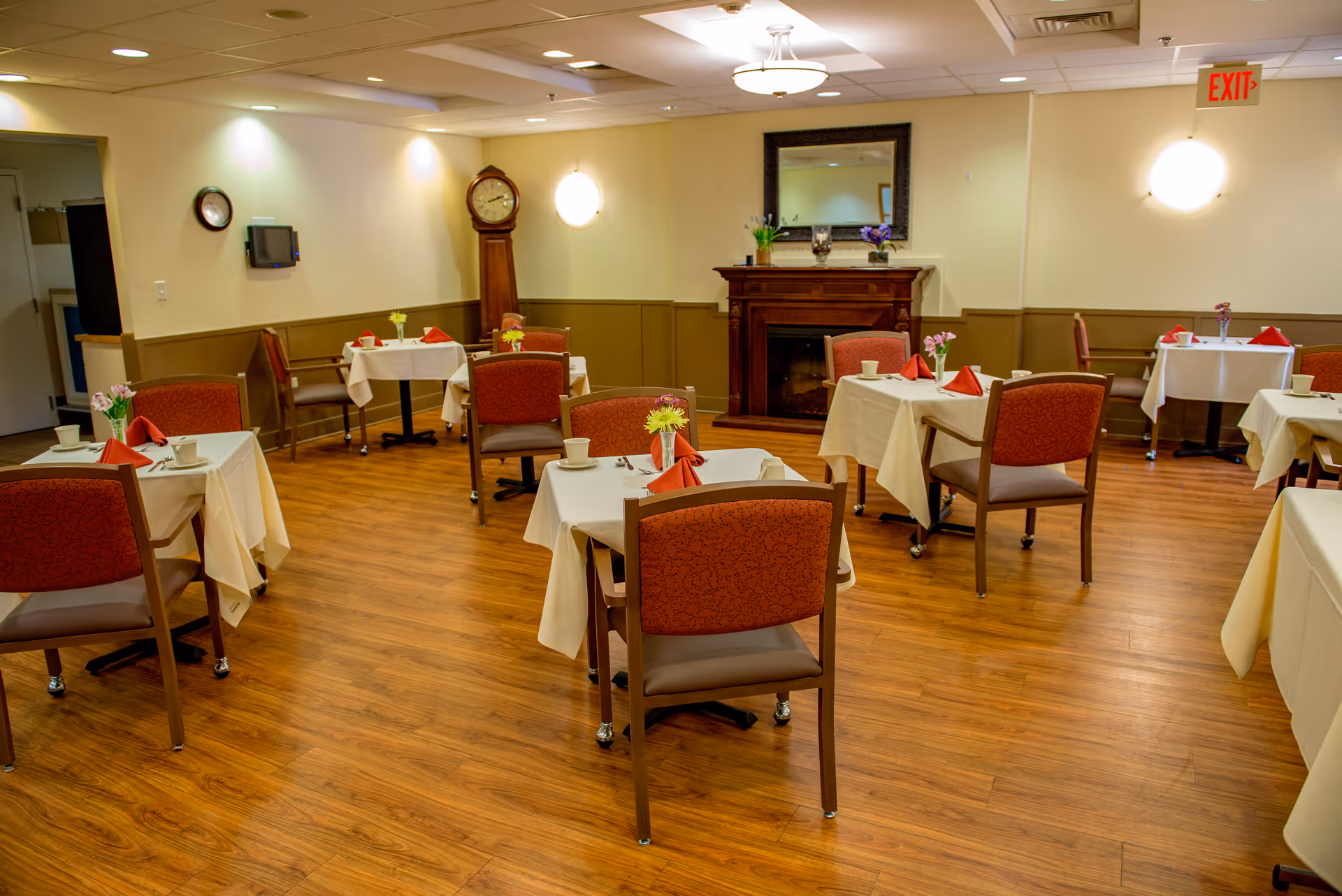 A dining room with several tables covered in white tablecloths, each set with cups, plates, and red folded napkins. The room has wooden flooring, red cushioned chairs, a grandfather clock, a fireplace with a mirror above it, and soft lighting. There are small flower arrangements on each table.
