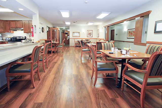 A dining area with wooden floors, multiple tables and chairs arranged for seating, and a counter with stools on the left side. The room is well-lit with ceiling lights and decorated with framed artwork on the walls.
