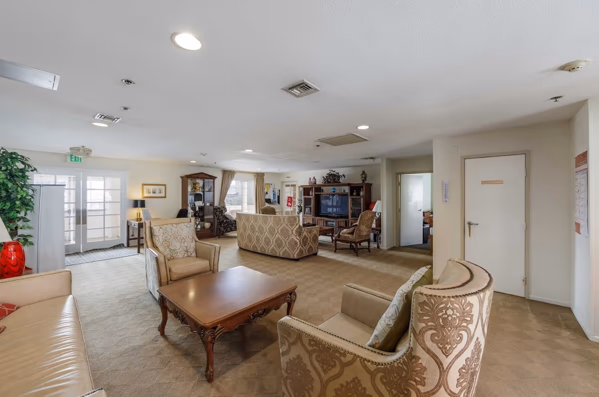 A spacious living room area in Westfield Villa Gardens featuring multiple upholstered armchairs and sofas arranged around a wooden coffee table. The room has beige carpeting, cream-colored walls, and recessed ceiling lights. There is a large wooden entertainment center with a television, decorative items, and a glass display cabinet. A door and windows with curtains are visible in the background, along with an exit door to the left.