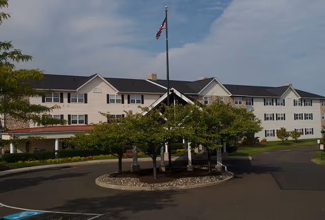 Front entrance of a three-story residential building with a covered drop-off, circular driveway, trees, and an American flag.