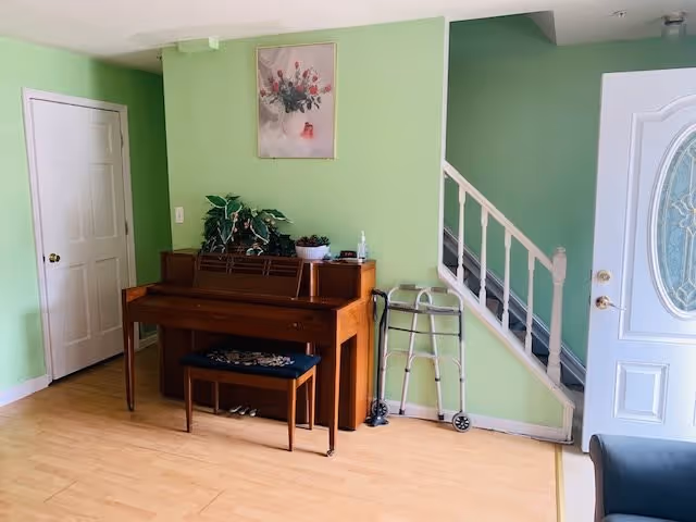 Interior view of a room with light green walls and wooden flooring. There is a brown upright piano with a matching bench, decorated with plants and a small bowl on top. A floral painting hangs above the piano. To the right of the piano is a walker and a staircase with white railing. A white door with a decorative glass panel is partially visible on the right side.