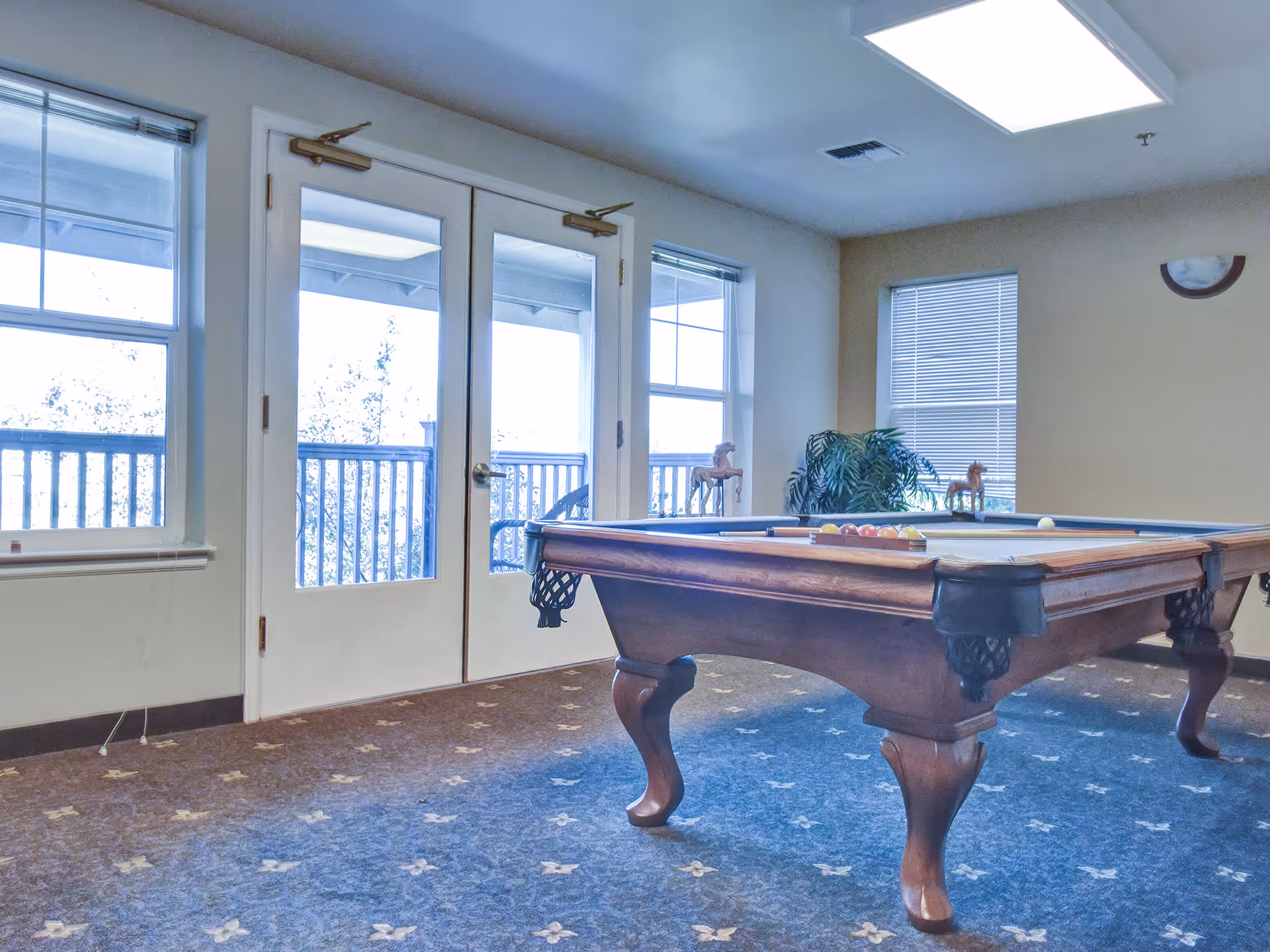 A room with a pool table set up for a game, featuring a wooden frame and legs. The room has carpeted flooring with a patterned design, two large windows, and a glass double door leading to an outdoor balcony with a railing. There is a potted plant and two small horse figurines on the windowsill. The ceiling has a rectangular light fixture.