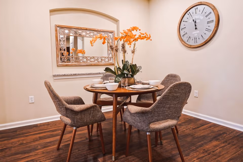 A small dining area with a round wooden table set for four, featuring white bowls and plates. Four upholstered chairs surround the table. A decorative mirror with a wooden frame hangs on the wall behind the table, and a large round wall clock with Roman numerals is mounted on the adjacent wall. The floor is dark wood, and the walls are painted a light beige color.