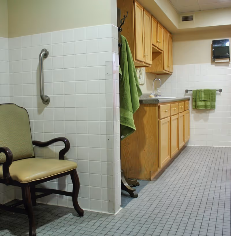 Interior view of a bathroom area with white tiled walls and floor, a wooden cabinet with a sink, green towels hanging on hooks and a towel rack, a grab bar on the wall, and a wooden chair with a cushioned seat and backrest.