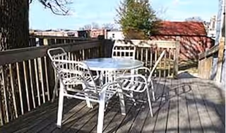 Outdoor wooden deck area with a round white metal table and four matching chairs, surrounded by a wooden railing and trees in the background.