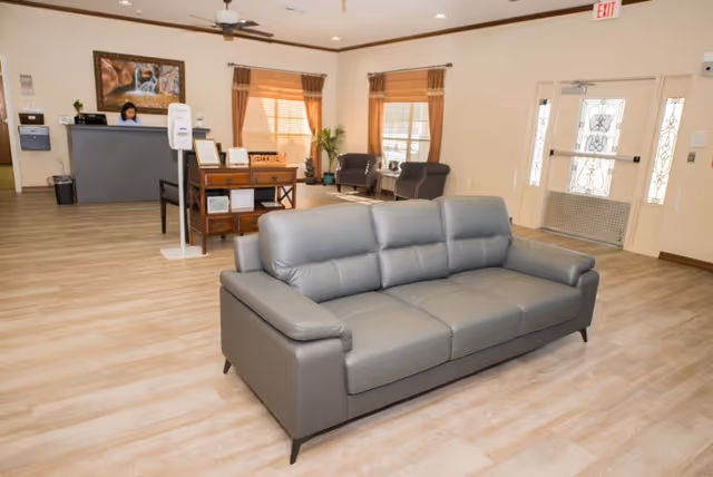 A spacious and well-lit reception area with a gray leather sofa in the foreground, a wooden desk with a welcome sign and hand sanitizer dispenser, a reception counter with a staff member behind it, two armchairs near windows with brown curtains, and a glass door with decorative metalwork.