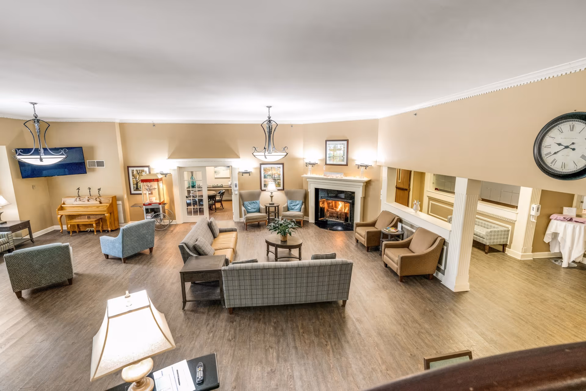 A spacious and well-lit living room area in a senior living facility named Shorehaven, featuring multiple armchairs and sofas arranged around a central coffee table with a plant. There is a fireplace with a fire burning, framed artwork on the walls, a large clock, a piano, a popcorn machine, and a flat-screen TV mounted on the wall. The room has wooden flooring and beige walls with white crown molding.