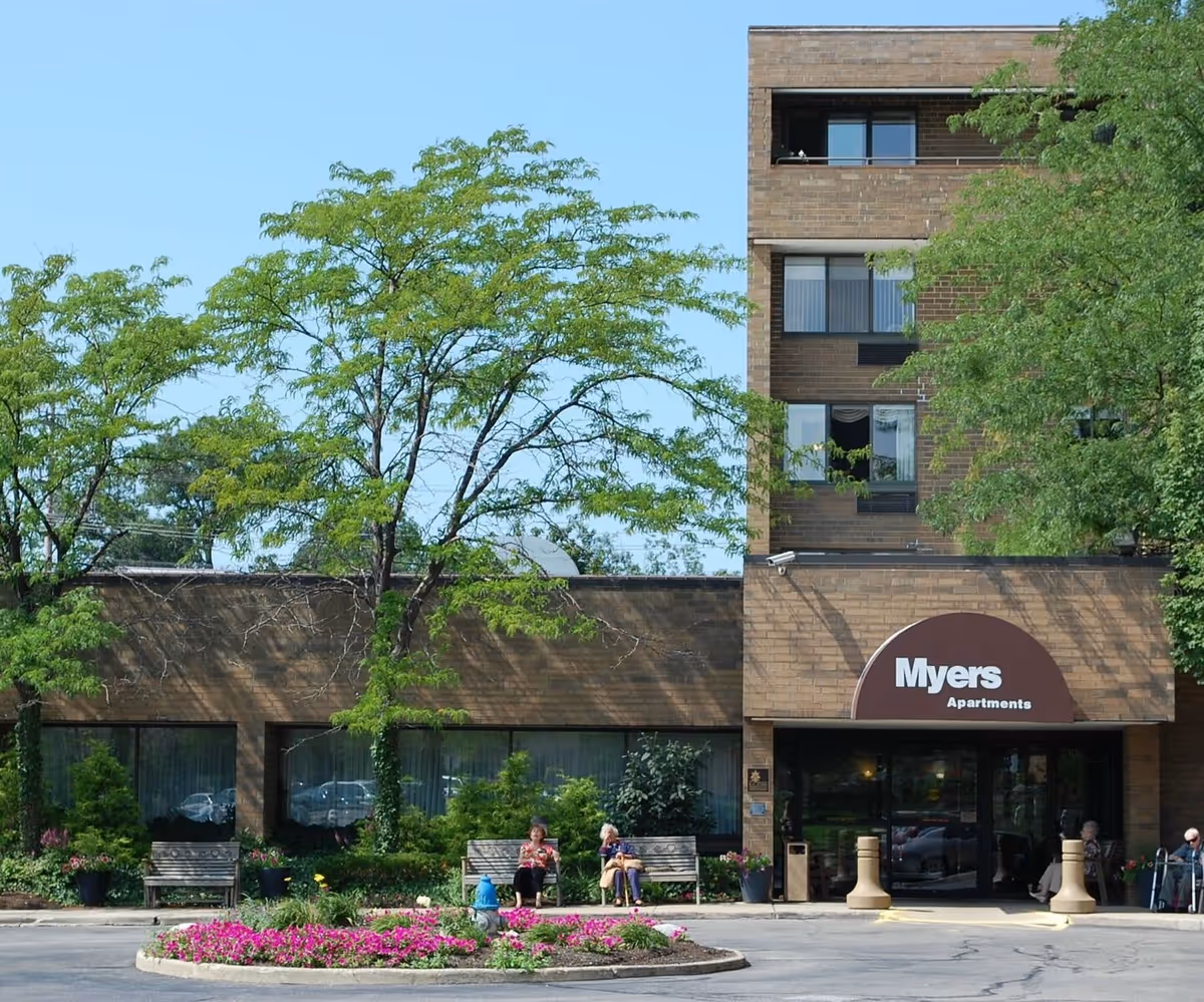 Front view of the Myers Apartments building showing the entrance awning, landscaping with a flower bed, benches and trees.