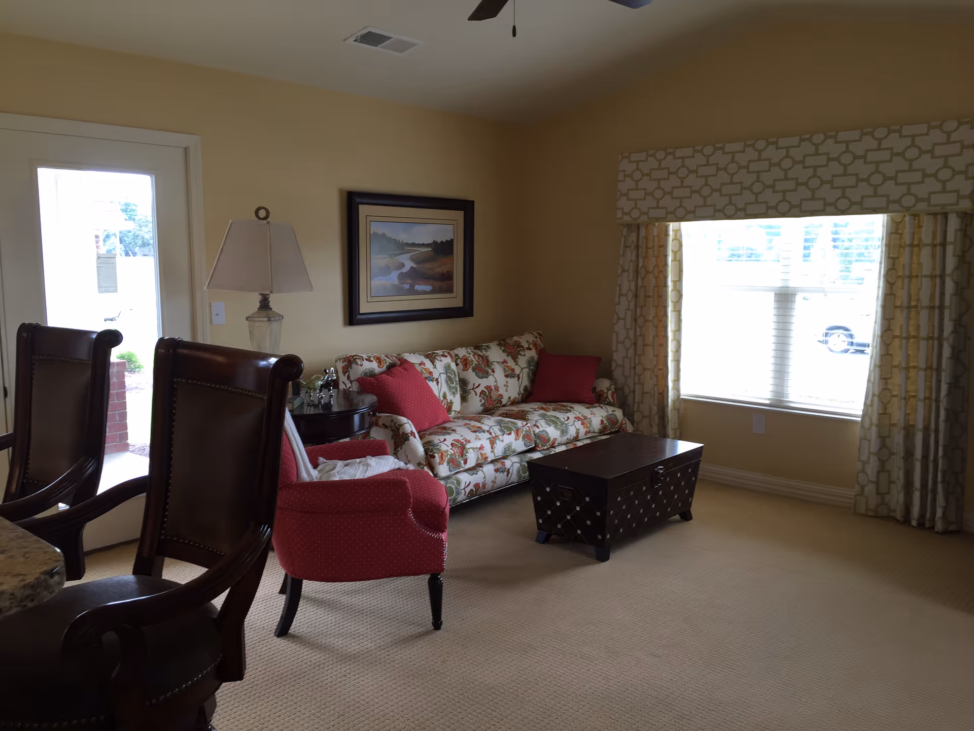 A cozy living room with a floral patterned sofa adorned with red cushions, a red armchair, a dark wooden coffee table, and two high-backed chairs near a counter. The room has beige walls, a large window with patterned curtains, a framed landscape painting, and a table lamp on a side table next to the sofa.