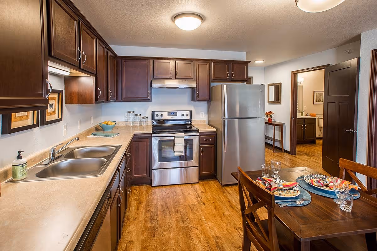 A modern kitchen with dark wood cabinets, stainless steel appliances including a refrigerator and stove, a double sink, and a wooden dining table set for four with plates, glasses, and napkins. The kitchen has wood flooring and a doorway leading to another room.