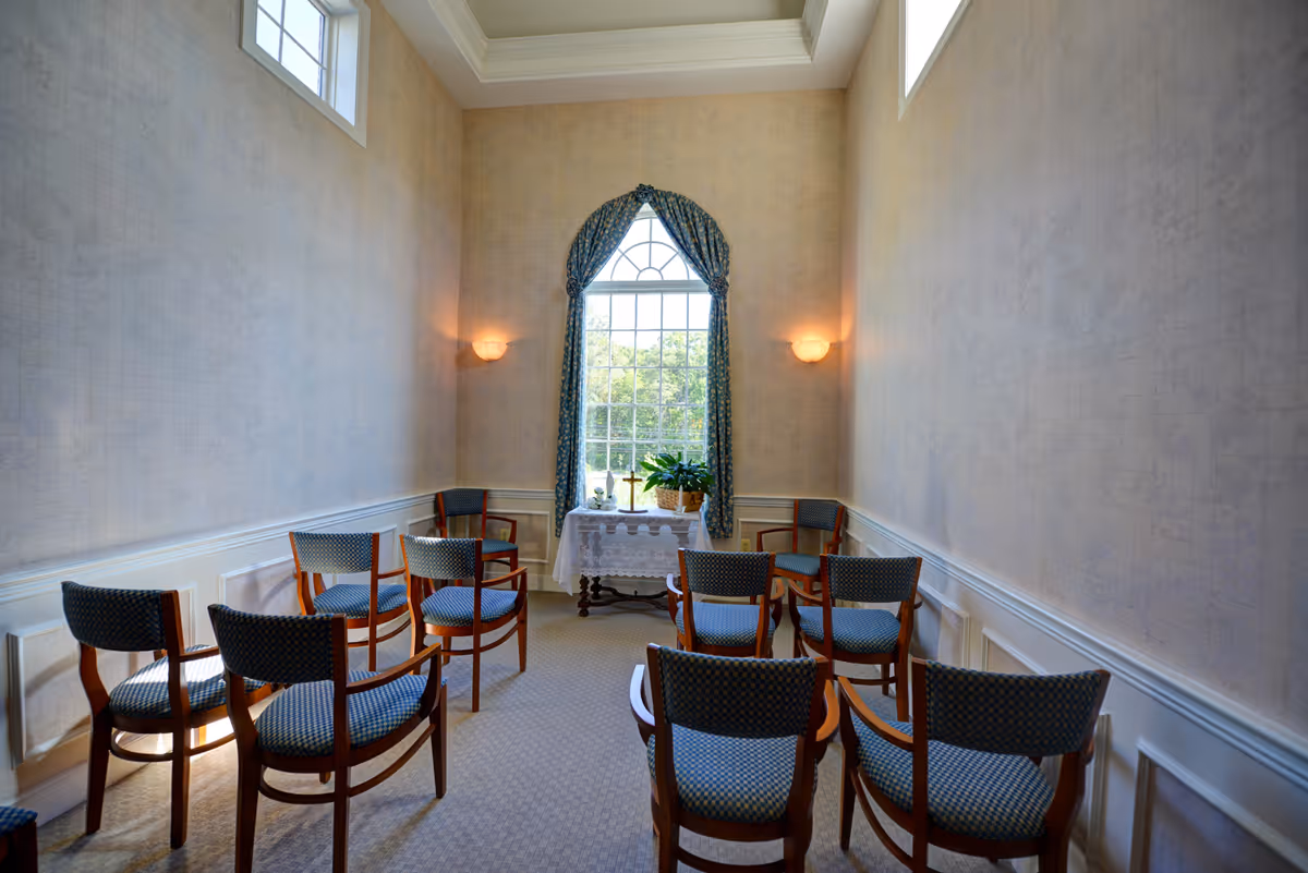 A small chapel or meditation room with wooden chairs arranged in rows facing a table covered with a white cloth, holding a plant and a cross. The room has high ceilings, beige walls, a large window with blue patterned curtains, and soft wall sconces providing warm lighting.