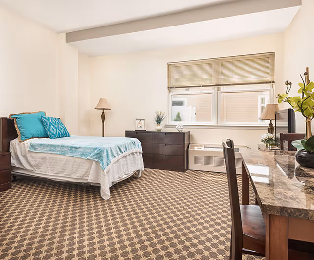 A bright and clean bedroom with a single bed covered in white and turquoise bedding, a patterned carpet, a dark wooden dresser with decorative items, a desk with a marble-like surface and a chair, two table lamps, and a large window with blinds letting in natural light.