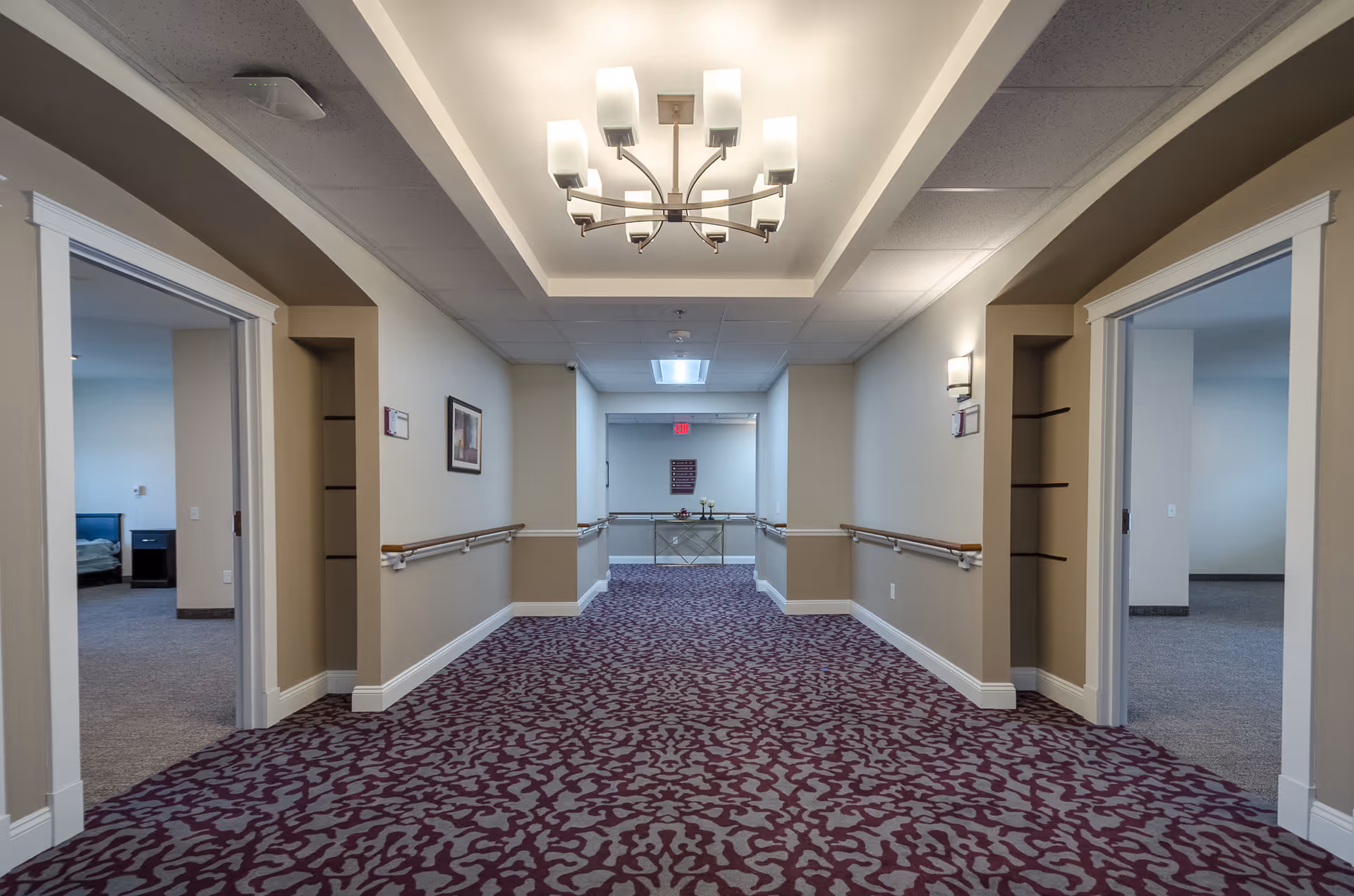 A carpeted hallway in a senior living facility with beige walls and white trim. The hallway has handrails on both sides and a modern chandelier hanging from the ceiling. Two open doorways on either side reveal rooms with beds and nightstands. At the end of the hallway, there is a small table with decorative items and a framed sign on the wall above it.
