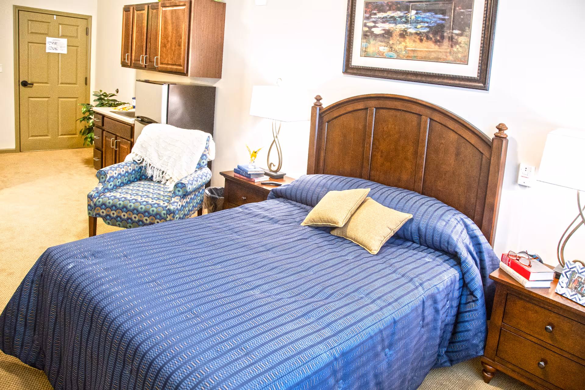A cozy bedroom with a large wooden bed covered in a blue bedspread and two beige pillows. There are two wooden nightstands on either side of the bed, each with a lamp. A framed painting hangs above the bed. To the left, there is a patterned armchair with a white throw draped over it, and a small kitchenette area with cabinets, a mini fridge, and a countertop. A closed door is visible in the background.