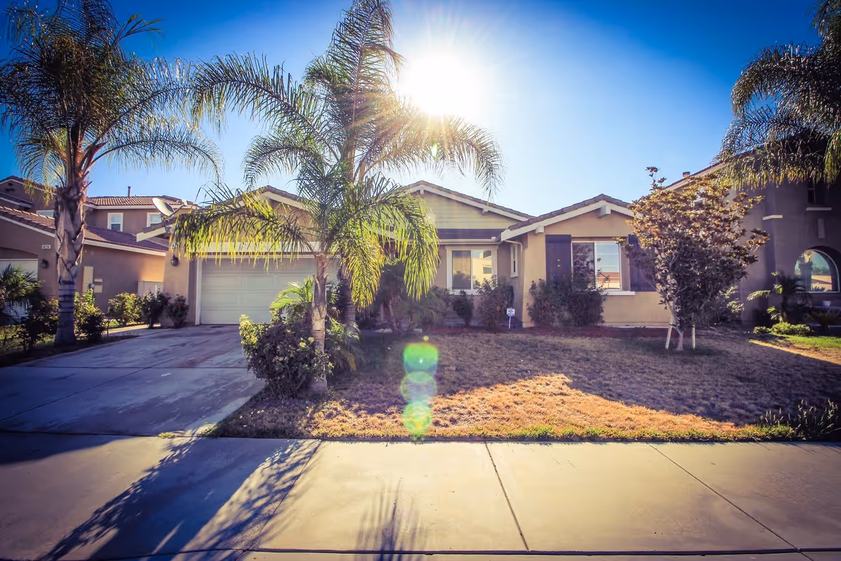 Front exterior view of a single-story residential building with a driveway, garage, palm trees, and a lawn under a bright sunny sky.