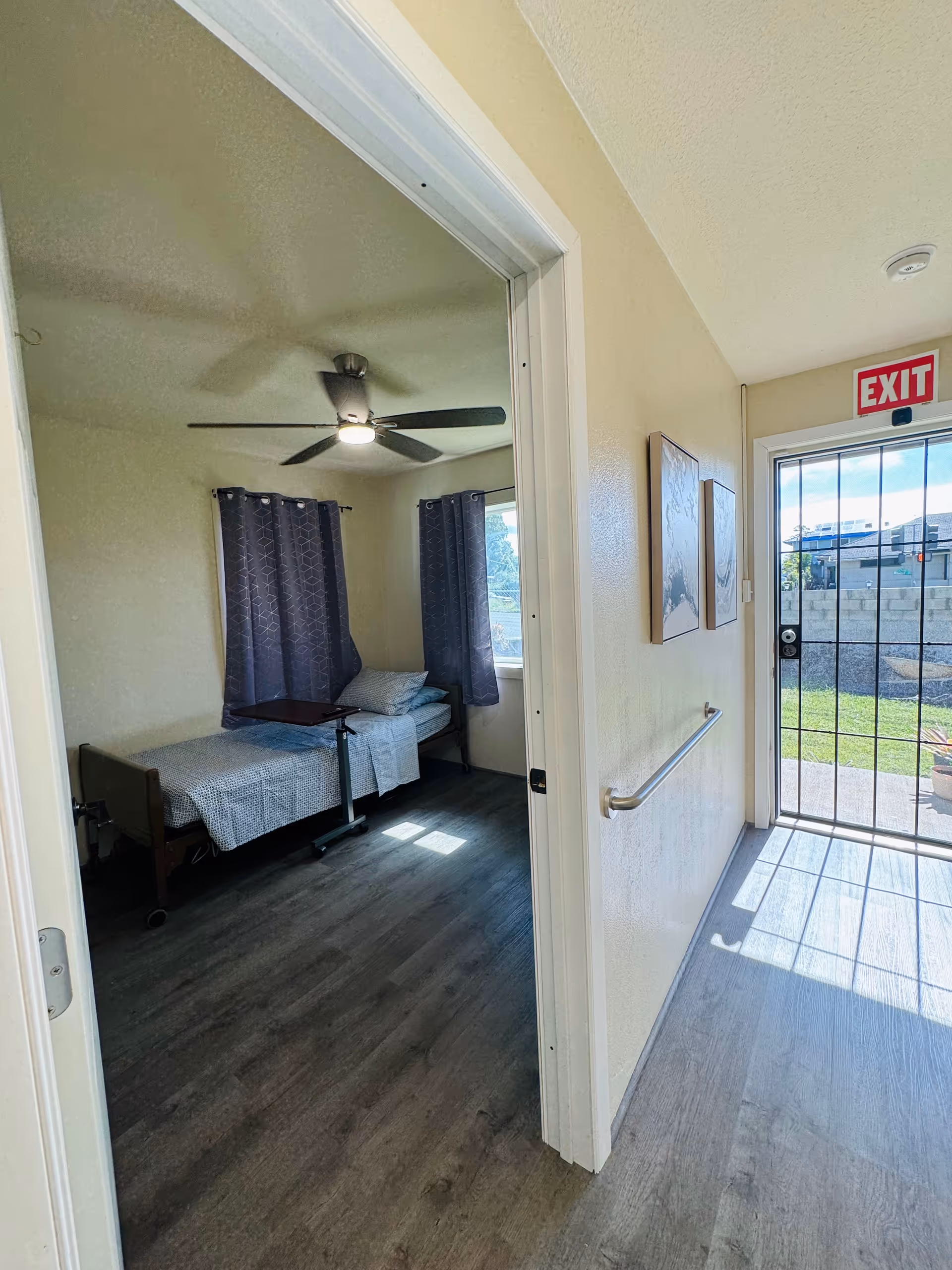 View down a hallway with a handrail on the right wall leading to a glass door with an exit sign above it. To the left, an open door reveals a small bedroom with a single bed, a bedside table, dark curtains on the windows, and a ceiling fan with a light.