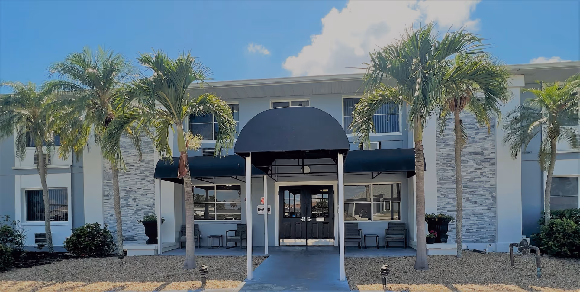 Front exterior view of a two-story building with a black awning over the entrance, flanked by palm trees and landscaping with gravel and bushes under a blue sky with some clouds.