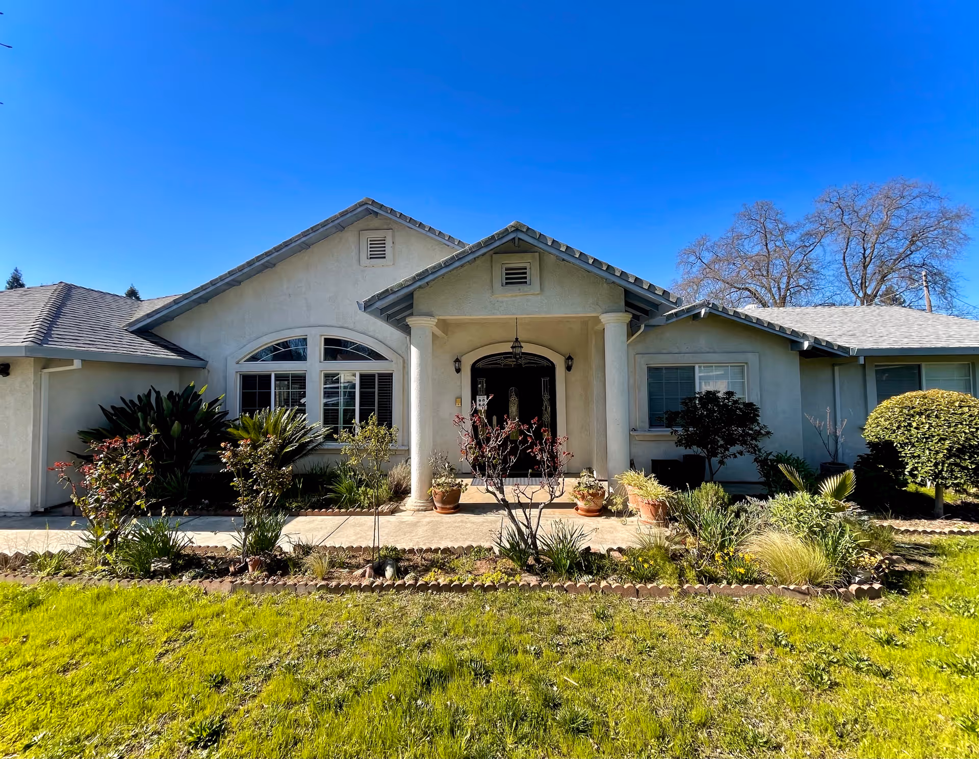 Front exterior of a single-story house with a covered entryway, columns, arched window and landscaped front lawn under a clear blue sky.