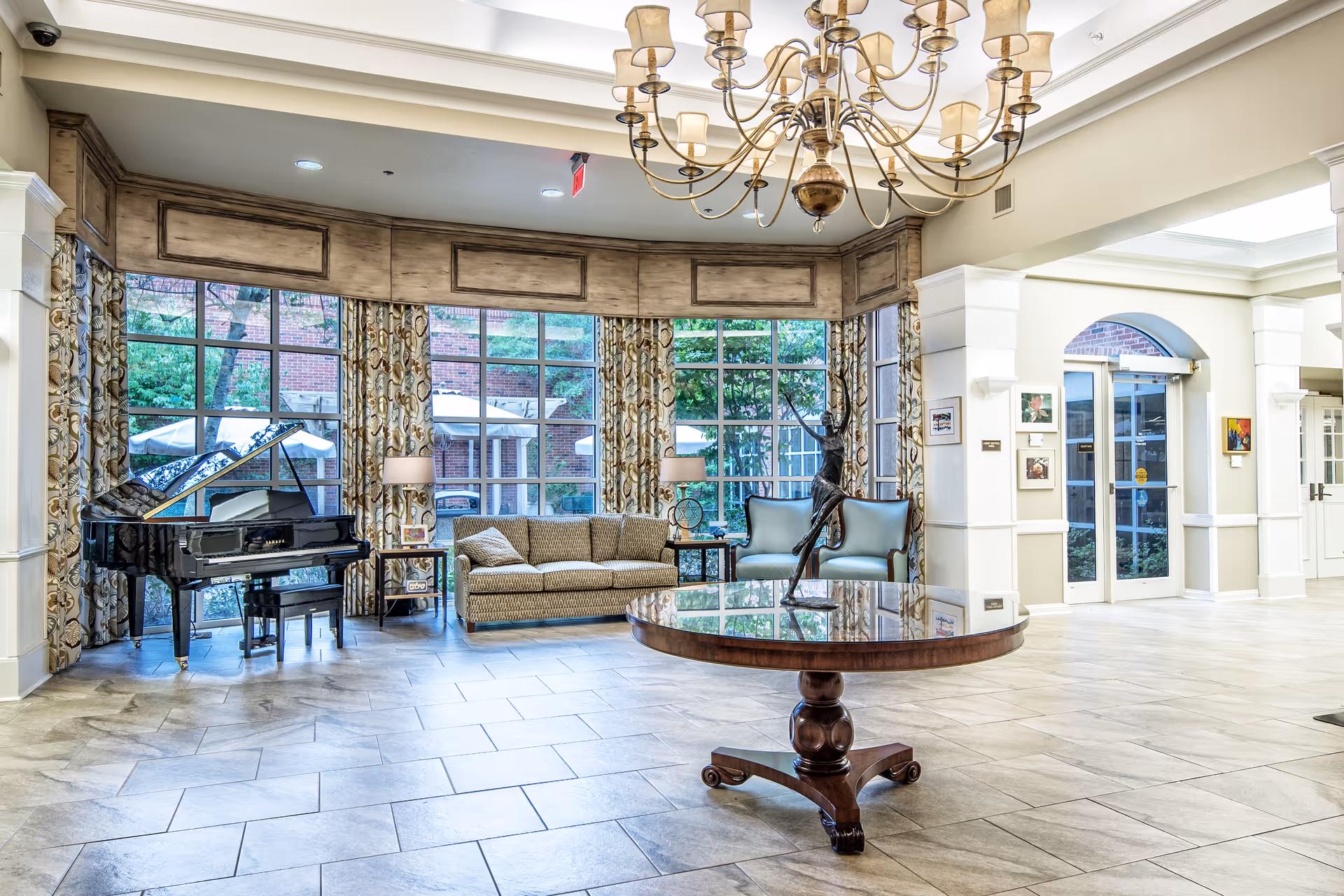 A spacious and well-lit common area with large windows covered by patterned curtains, a grand piano on the left, a sofa and two armchairs near the windows, and a round wooden table with a decorative sculpture in the center. The floor is tiled, and a chandelier hangs from the ceiling. There are double glass doors on the right side leading outside or to another area.