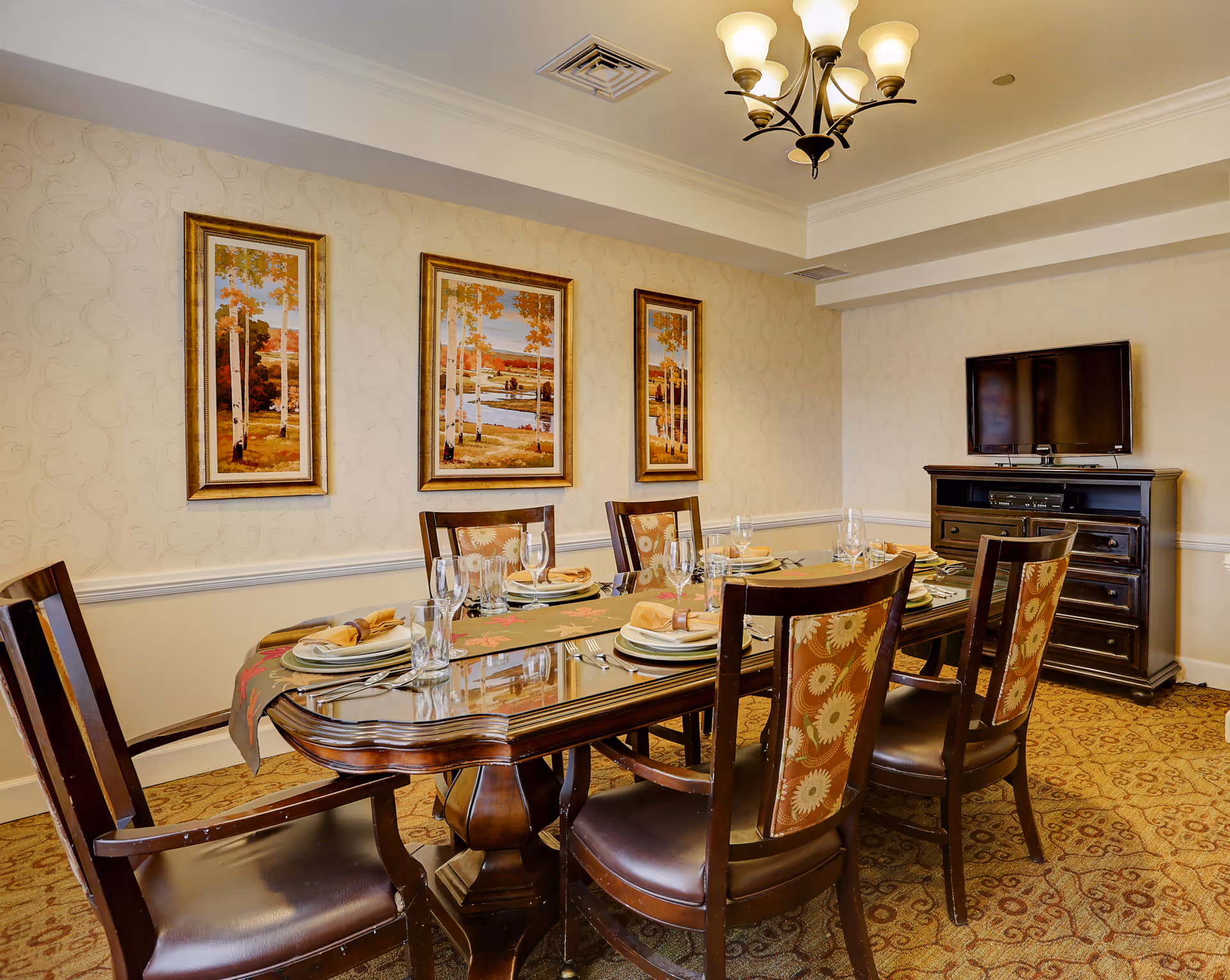 A dining room with a wooden table set for six people. The table has plates, glasses, napkins, and silverware arranged neatly. There are six chairs with patterned upholstery around the table. On the wall behind the table are three framed paintings depicting autumn trees and a river. A dark wooden cabinet with a flat-screen TV on top is positioned against the adjacent wall. The room has beige wallpaper with a subtle pattern and a chandelier with five lights hanging from the ceiling.