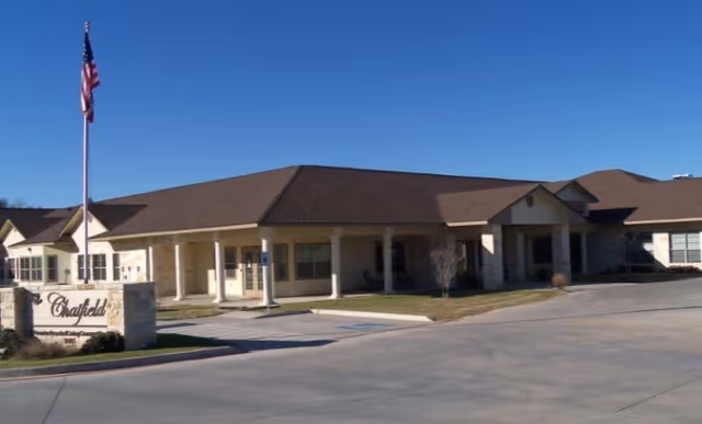 Exterior view of The Chatfield Assisted Living facility showing a single-story building with a brown roof, beige walls, a covered entrance supported by columns, an American flag on a flagpole, and a stone sign with the facility's name.