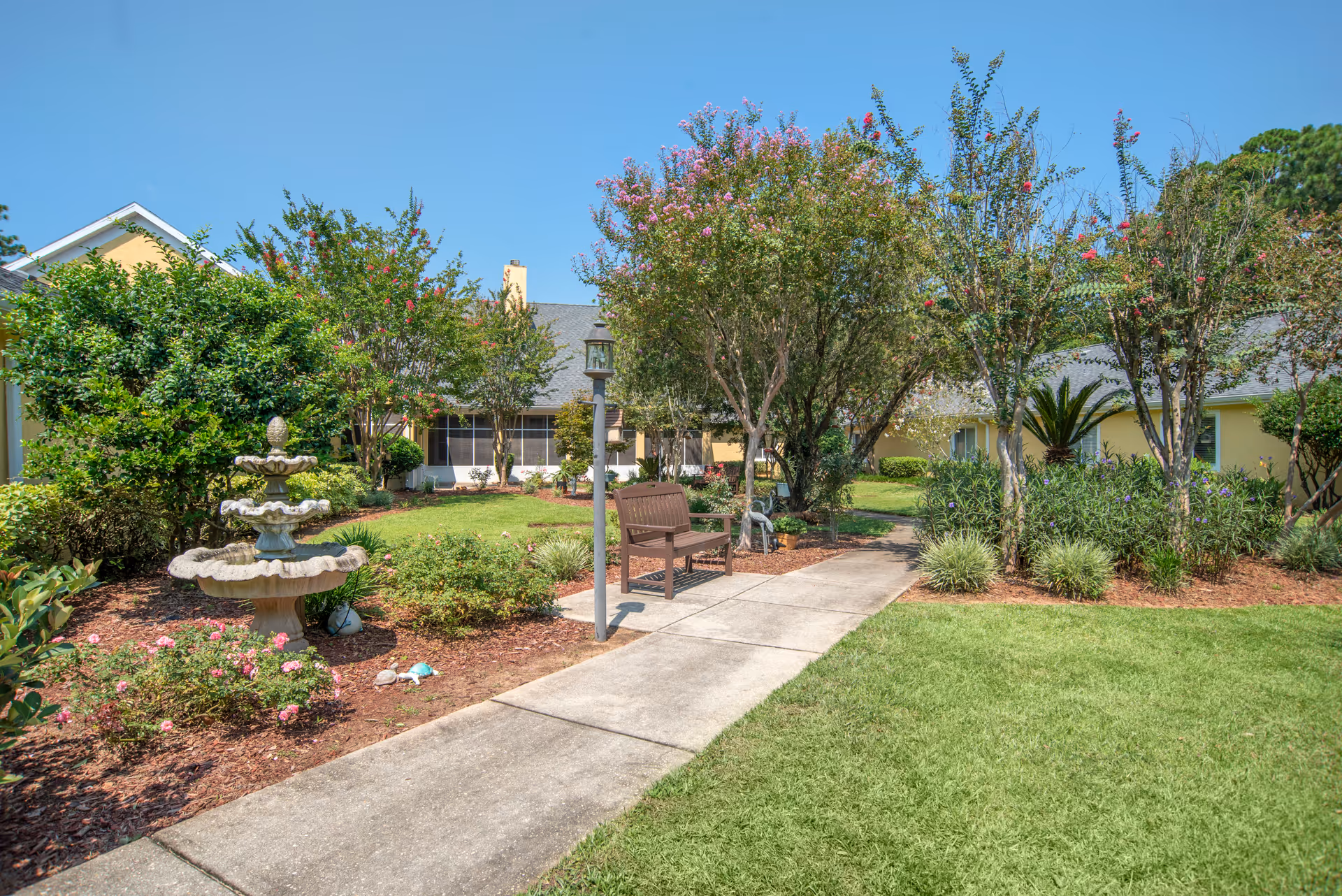 A peaceful outdoor garden area at Brookdale Pensacola featuring a concrete walkway, a wooden bench, a multi-tiered stone fountain, various flowering bushes, and trees with pink blossoms under a clear blue sky.