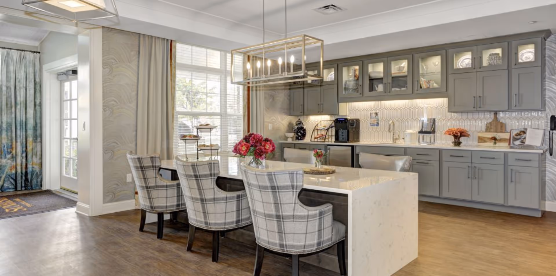 Bright communal dining area and kitchenette with a marble island, plaid upholstered chairs, gray cabinets, and decorative lighting.