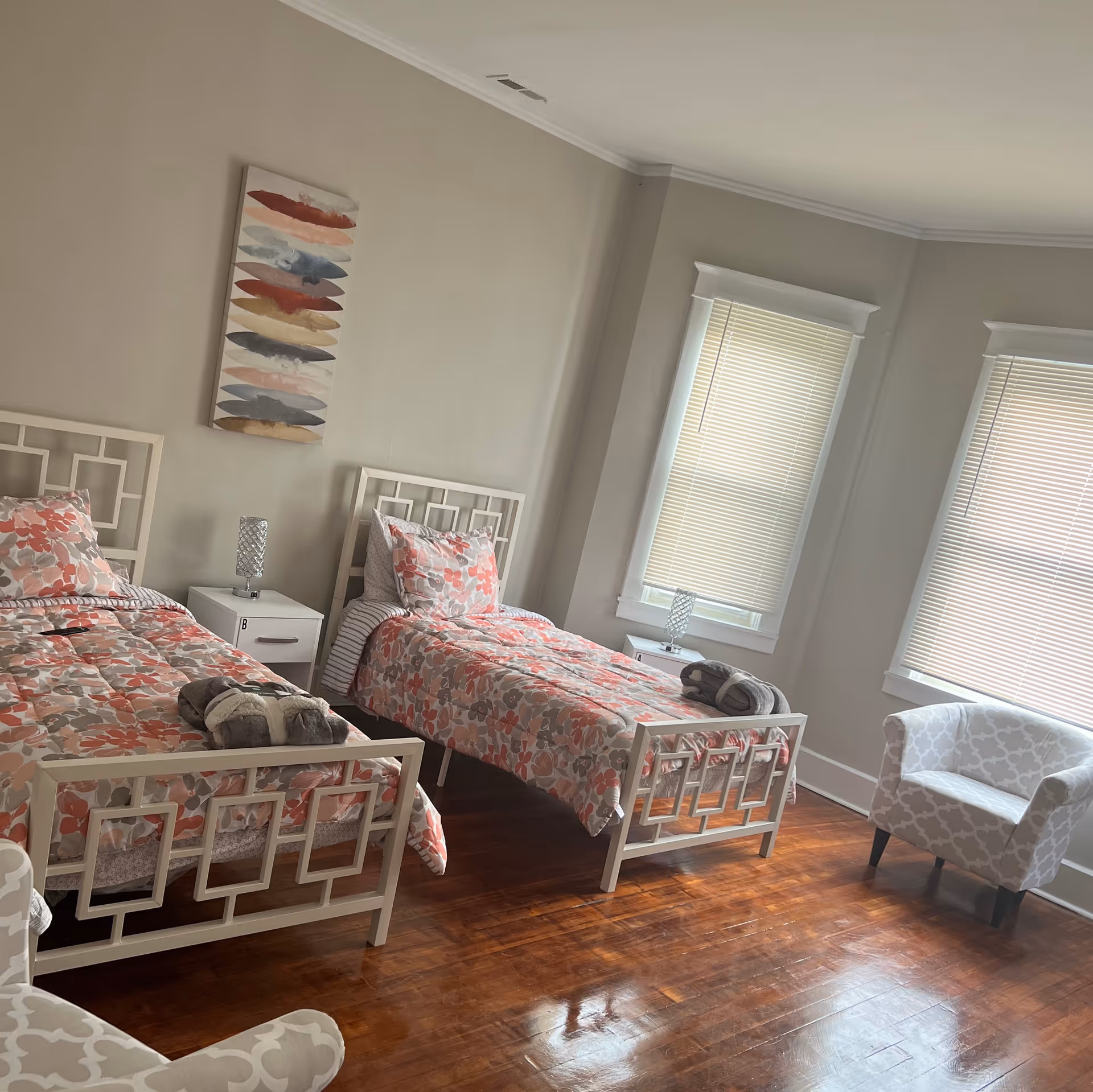 Sunlit bedroom with two twin beds dressed in floral bedding, a shared nightstand, and two windows with blinds.
