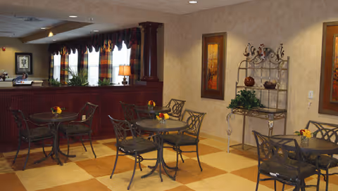 Interior view of a dining area with several metal tables and chairs arranged on a checkered tile floor. The room features warm beige walls with framed artwork and a decorative metal shelving unit holding plants and ornaments. A counter with a dark wood finish and windows with valances is visible in the background, along with ceiling lights providing illumination.