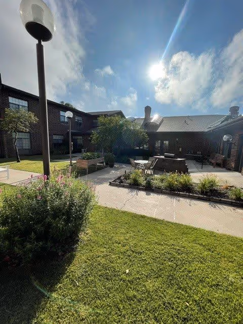 Outdoor courtyard area of a senior living facility with green grass, flower beds, benches, tables with chairs, and lamp posts. The building surrounds the courtyard and the sun is shining brightly in a partly cloudy sky.