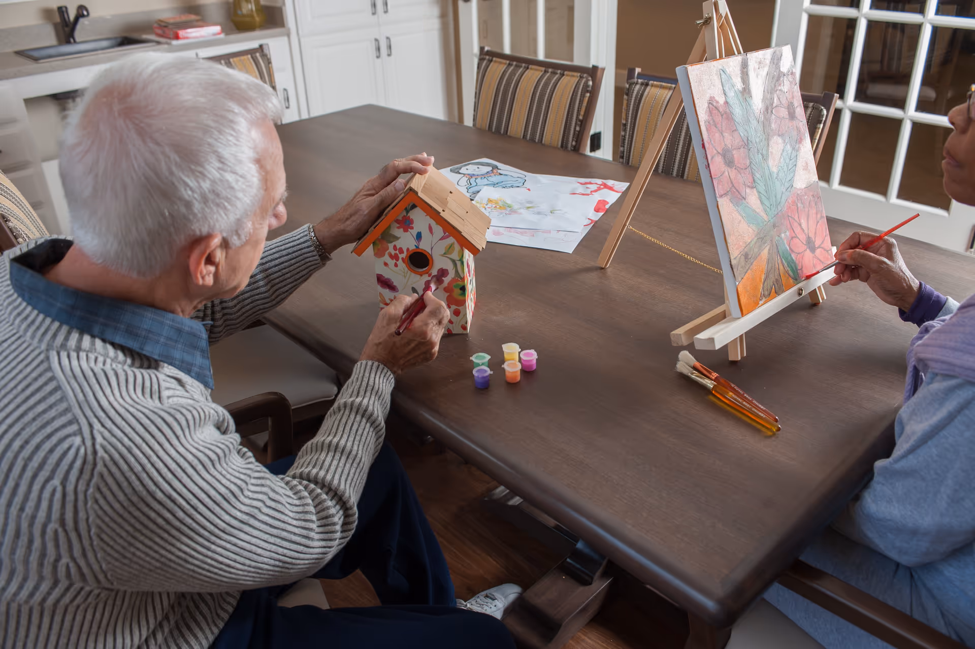 Two elderly individuals sitting at a wooden table engaged in painting activities. One person is painting a small birdhouse with floral designs, while the other is painting a canvas on an easel with a floral and leaf design. Various paint pots and brushes are on the table.