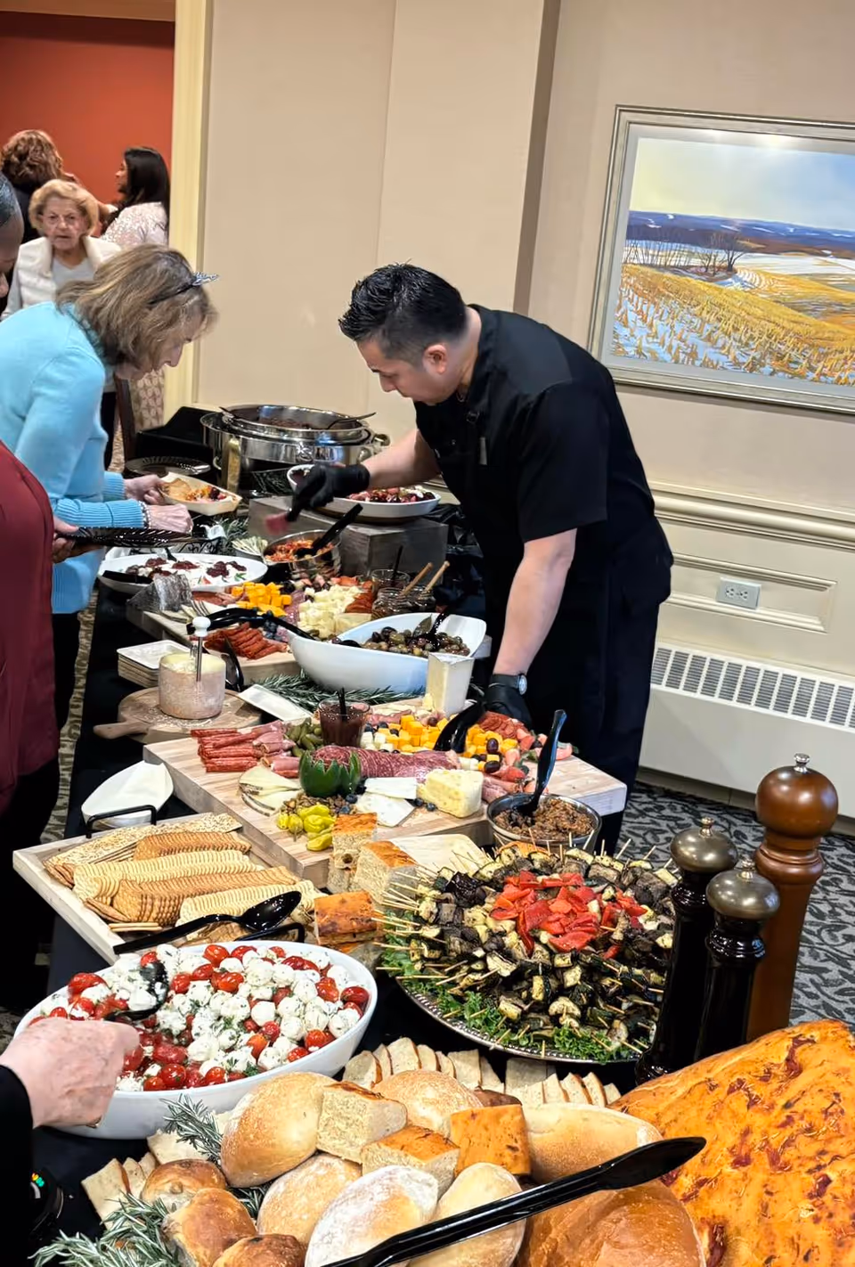 A buffet table filled with various foods including bread rolls, crackers, cheese, skewers with vegetables and meat, and a bowl of mozzarella and cherry tomato salad. A man wearing black gloves and a black shirt is serving food while several elderly people stand nearby, selecting their food. The setting appears to be indoors with a framed landscape painting on the wall.