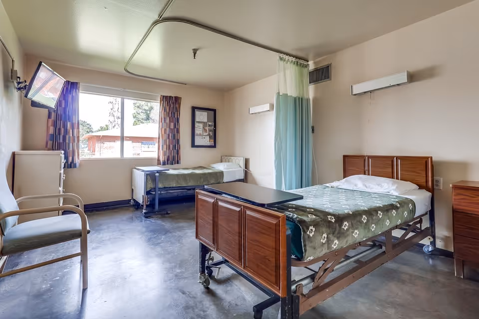 A nursing home bedroom with two hospital-style beds, each with green floral blankets. There is a chair near the left wall, a window with patterned curtains, a wall-mounted TV, and a privacy curtain between the beds. The room has a polished concrete floor and neutral-colored walls.