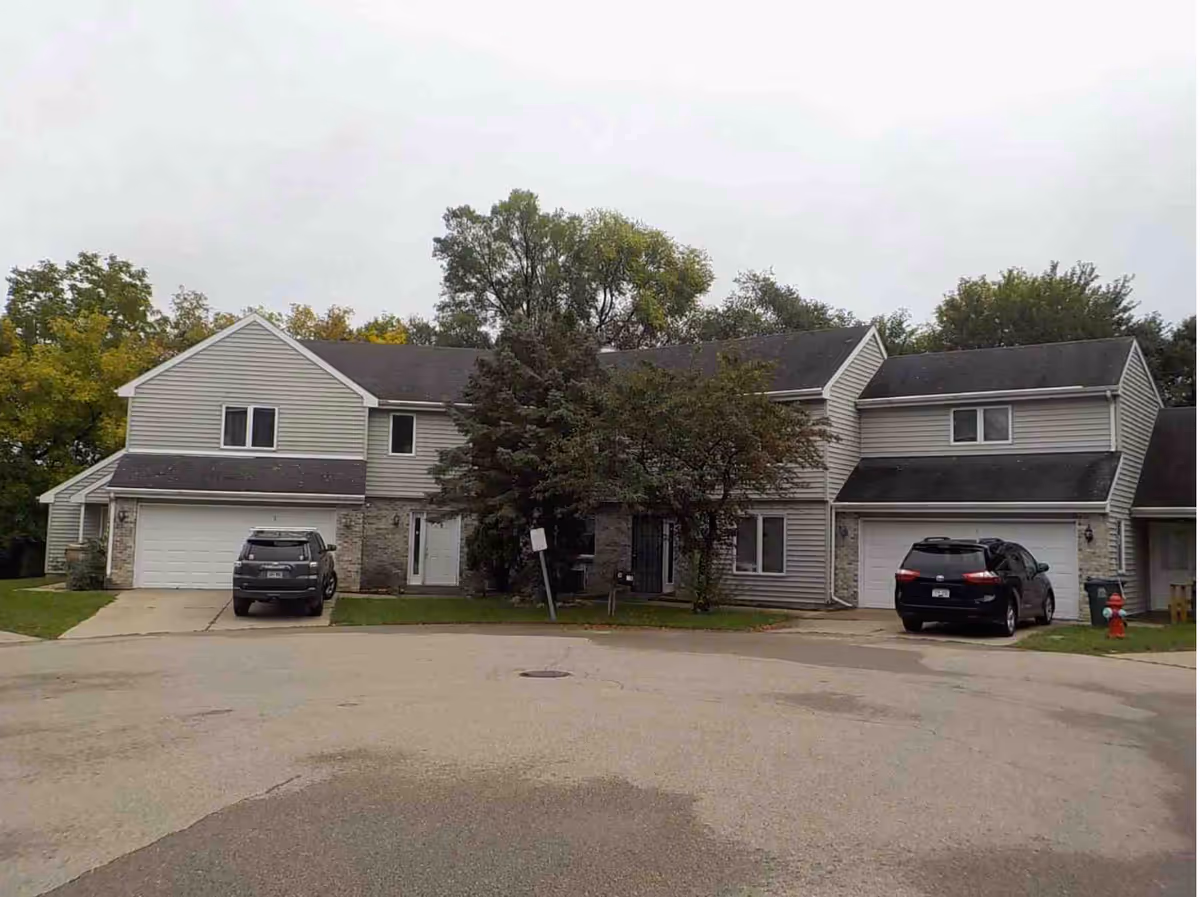 Two-story townhouse-style building with attached garages, parked cars, and trees in front under an overcast sky.
