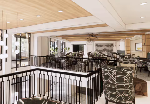 Spacious and modern common area in a senior living facility with patterned armchairs, multiple dining tables and chairs, large windows letting in natural light, decorative ceiling beams, and contemporary light fixtures.