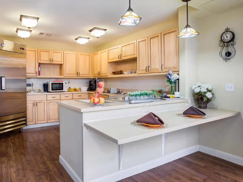 A clean and well-lit kitchen area with light wood cabinets, a stainless steel refrigerator, a microwave, and a kitchen island with a built-in sink. The island has a breakfast bar with two place settings, each with a brown napkin and a glass. There are decorative flowers on the counter and a wall clock on the right wall.