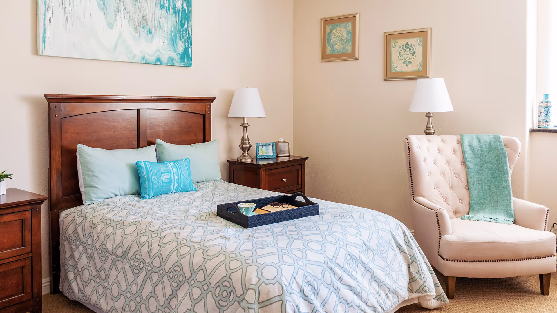 A cozy bedroom featuring a wooden bed with a patterned bedspread and multiple pillows in shades of blue and green. There are two wooden nightstands with lamps on either side of the bed. A beige upholstered armchair with a green throw blanket is positioned near a window, and two framed artworks hang on the wall above the chair.
