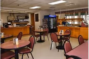 Interior view of a dining area in a senior living facility with round tables and chairs. The room features a food service counter, a refrigerated beverage display, and a buffet station. The ceiling has fluorescent lighting, and windows with valances allow natural light into the space.