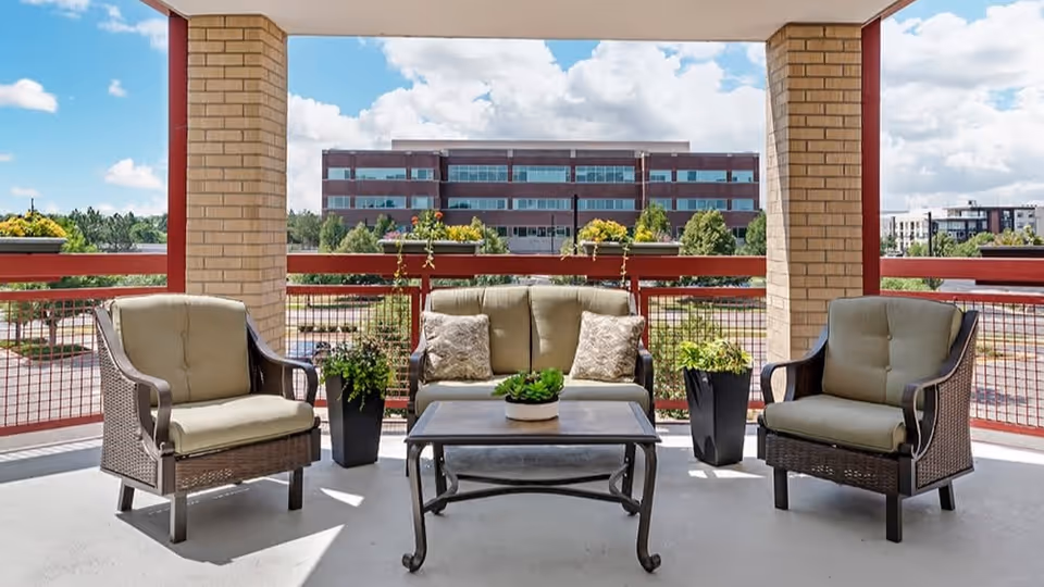 Outdoor covered patio area with cushioned wicker seating including two armchairs and a loveseat around a metal coffee table, with potted plants on either side and a view of a multi-story building and trees in the background under a partly cloudy sky.