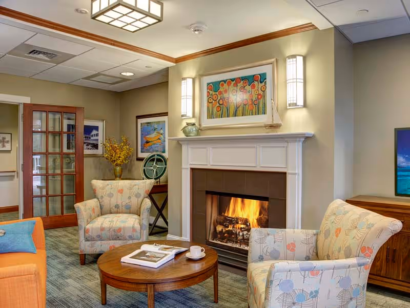 Cozy common room with a lit fireplace, patterned armchairs, a round coffee table and colorful artwork on the walls.