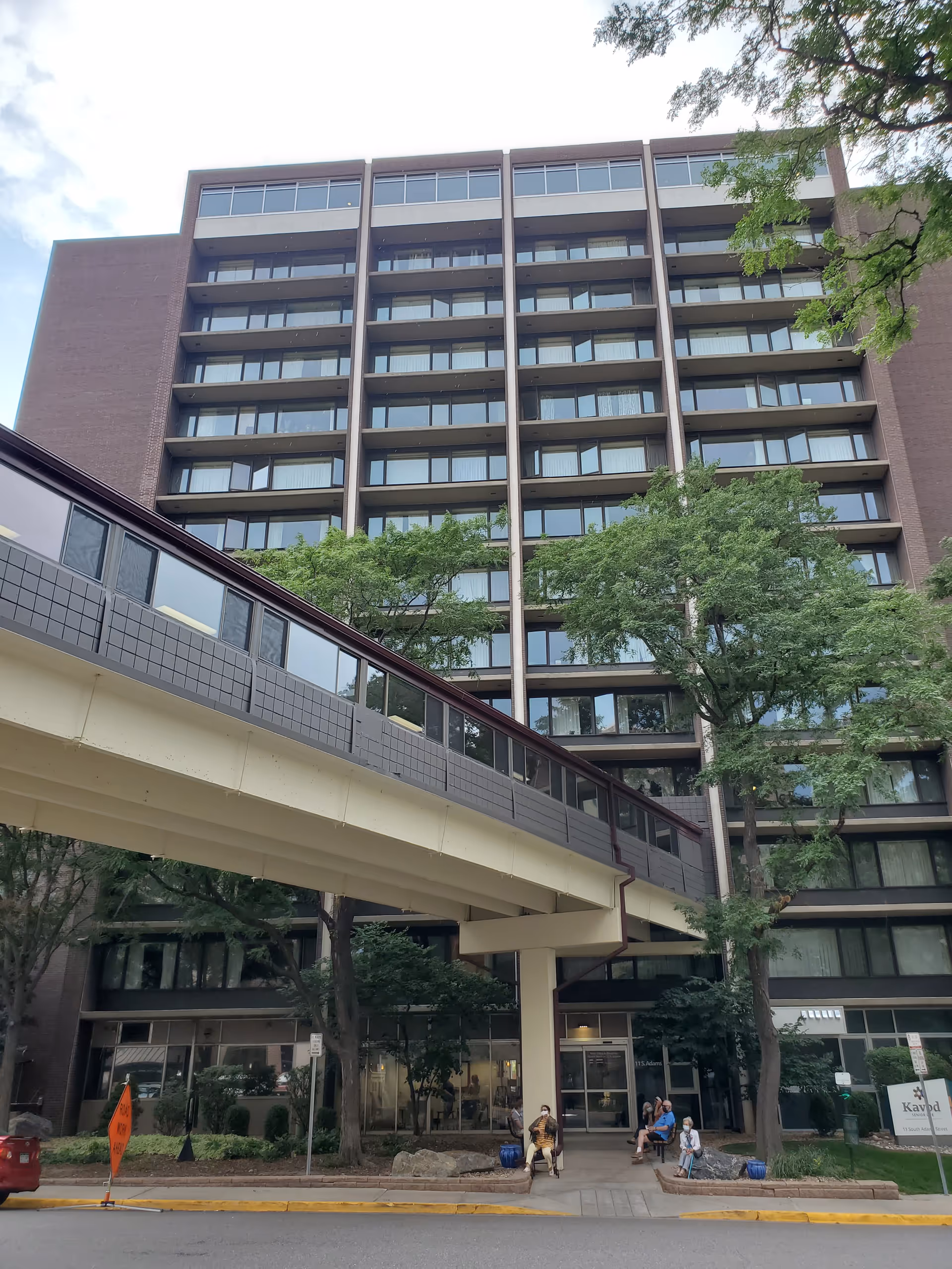 Exterior view of a multi-story senior living building with a covered pedestrian skywalk, trees, and people near the entrance.
