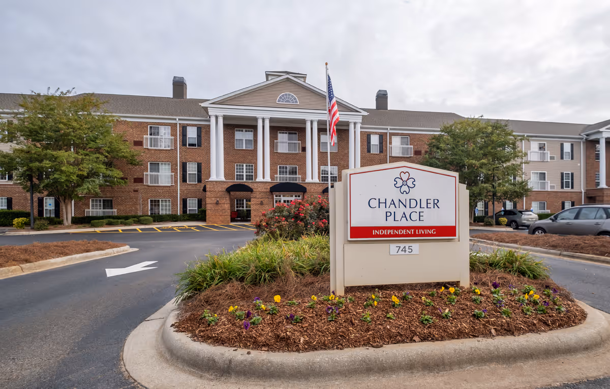 Front exterior view of Chandler Place Independent Living facility with a large sign in the foreground surrounded by flowers and landscaping. The building is a multi-story brick structure with white columns, balconies, and an American flag flying near the entrance.