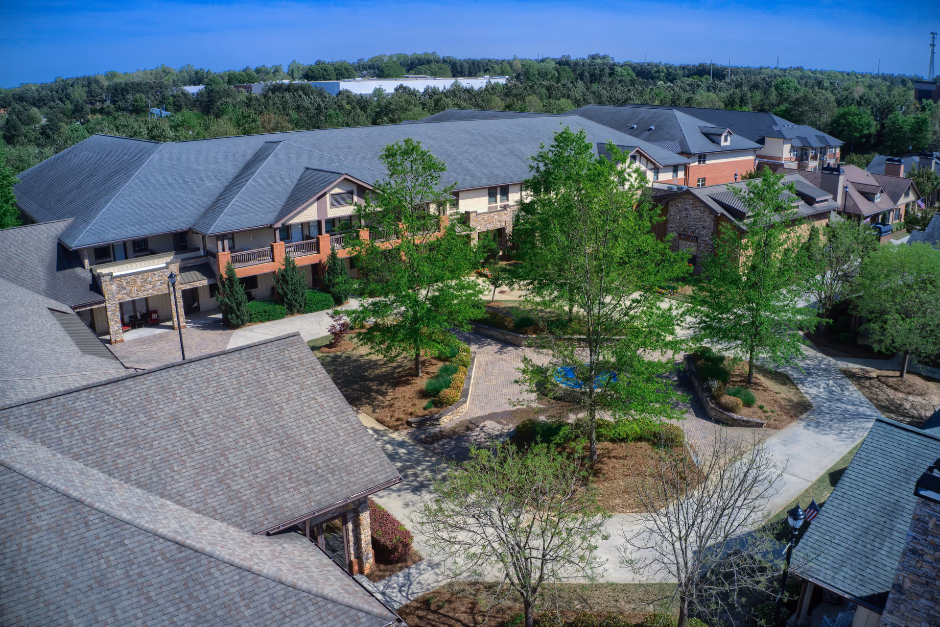 Aerial view of TerraBella Sugarloaf senior living facility showing multiple connected buildings with gray roofs surrounding a landscaped courtyard with trees and walking paths under a clear blue sky.