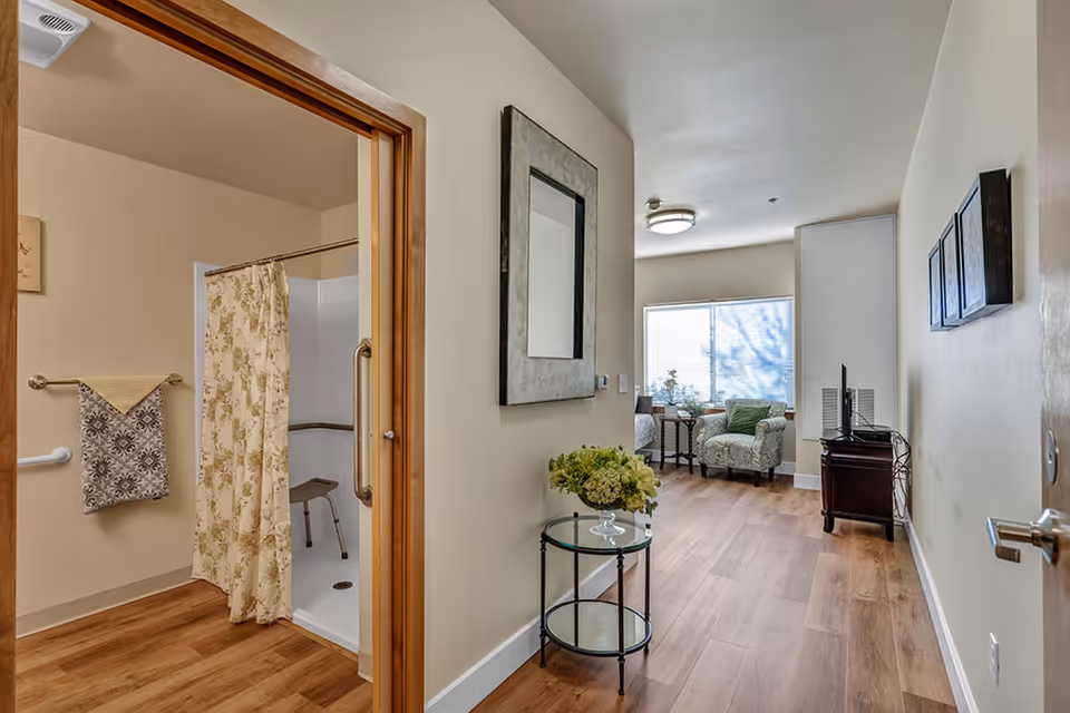View of a hallway in a senior living facility with a bathroom on the left featuring a shower with a floral curtain and grab bars. The hallway leads to a sitting area with two armchairs, a small table, and a window letting in natural light. The walls are light-colored and the floor is wood.